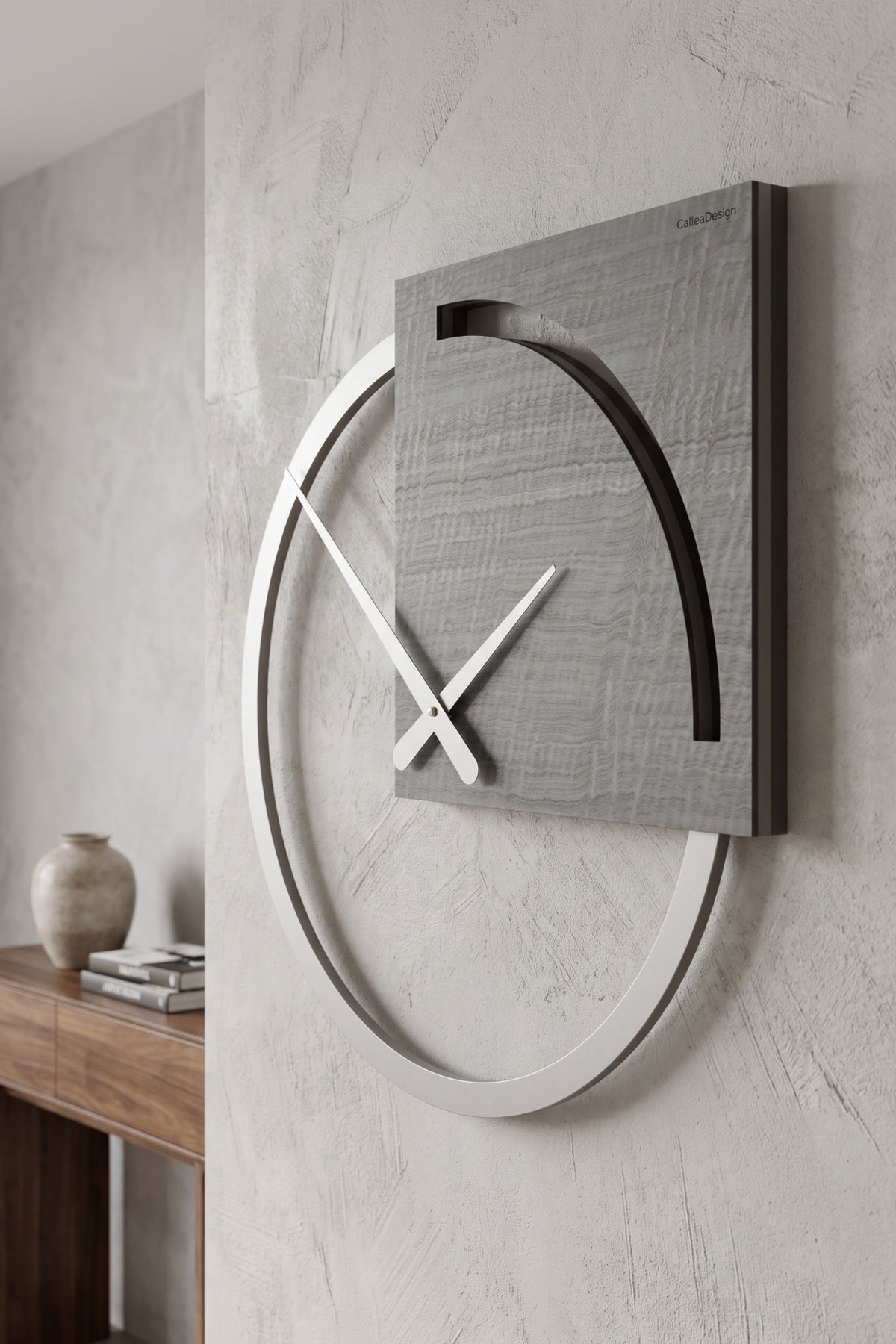 CalleaDesign Karl wall clock in Grey Burl finish shown from an angled close-up on a textured wall in a contemporary interior